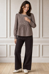Model wearing the Aspiga Mina Gingham Check Blouse in Chocolate/White with dark trousers, full-length front view, hands in pockets, smiling