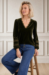 Model wearing the Aspiga Claudette Velvet Blouse in Dark Olive, seated on a stool, front view