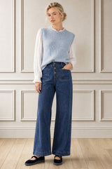 A woman stands in a room with paneled cream walls and light wood flooring, wearing a light blue knit sweater vest over a white long-sleeve blouse, dark blue wide-leg jeans, and black flats.