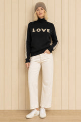 Model wearing the Aspiga Merino Wool Love Polo Neck Jumper in Black/Taupe with a taupe beanie and cream trousers, full-length front view