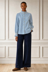 Model wearing the Aspiga Embroidered Smocked Baby Cord Blouse in Ice Blue tucked into navy trousers with brown loafers, full-length front view