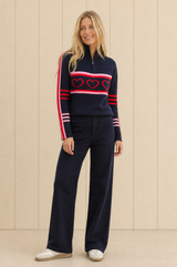 Model wearing the Aspiga Merino Wool Stripe Hearts Quarter Zip in Navy/Red/Light Pink with navy trousers, full-length front view, smiling