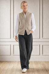Model wearing the Aspiga Merino Wool Zip Through Gilet in Taupe over a white blouse with black trousers, full-length front view, walking