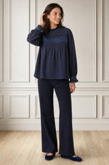 Model wearing the Aspiga Seersucker Embroidered Blouse in Navy with navy trousers, full-length front view, hand in hair, smiling