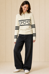 Model wearing the Aspiga Merino Wool Stripe Hearts Quarter Zip in Off White/Black with black jeans, full-length front view, smiling