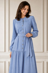 Model wearing the Aspiga Baby Cord Midi Dress in Ice Blue, a light blue corduroy belted shirt dress with long sleeves and a collar, alternate view