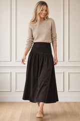 Model wearing the Aspiga Dropped Waist Baby Cord Skirt in Chocolate with a beige jumper, full-length front view, smiling
