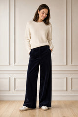 Model wearing the Aspiga Merino Wool Crew Neck Jumper in Ivory with navy corduroy trousers and white trainers, full-length front view, smiling
