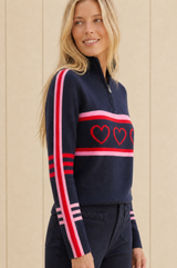Model wearing the Aspiga Merino Wool Stripe Hearts Quarter Zip in Navy/Red/Light Pink with navy trousers, side view, smiling
