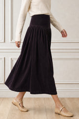 Model wearing the Aspiga Dropped Waist Baby Cord Skirt in Aubergine with a cream jumper, side view walking