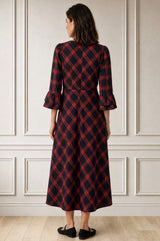 Model wearing the Aspiga Elizabeth Check Dress in Red/Blue with black shoes, full-length front view outdoors among stone columns, smiling