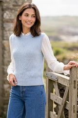 A woman wearing a light blue ribbed knit sweater vest over a white long-sleeve blouse and blue jeans, standing outdoors next to a weathered wooden fence.