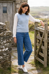 A woman wearing a light blue ribbed knit sweater vest over a white long-sleeve top, blue wide-leg jeans, and white sneakers, standing outdoors next to a rustic wooden gate.