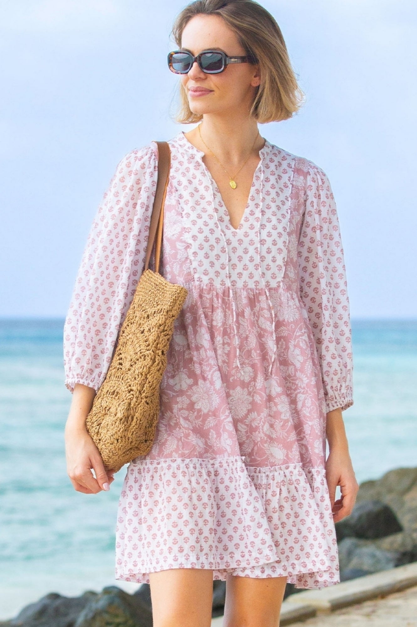 Woman in a pink floral dress standing on a beach with sunglasses and a raffia bag.