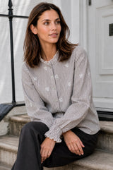 Model wearing the Aspiga Cosima Embroidered Blouse in Chocolate/White with dark trousers, seated on stone steps near a white door