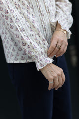 Close-up of a model wearing the Aspiga Vienna Printed Long Sleeve Blouse in Buta Pink/Cream, hand near collar, outdoors by railings