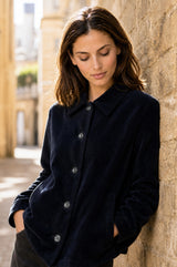 Model wearing the Aspiga Corduroy Collared Jacket in Deep Navy, leaning against a sunlit stone wall, buttoned up, looking down