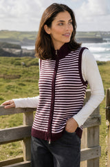 Model wearing the Aspiga Merino Wool Stripe Zip Through Gilet in Burgundy/Pink, leaning against a wooden fence outdoors by the coast