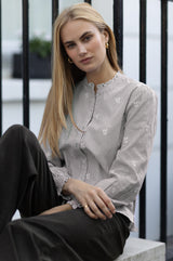 Model wearing the Aspiga Cosima Embroidered Blouse in Chocolate/White with dark trousers, seated on outdoor steps beside a railing
