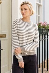 Model wearing the Aspiga Merino Wool Button Through Stripe Cardigan in Taupe/Cream with black trousers, full-length view outdoors, looking to the side