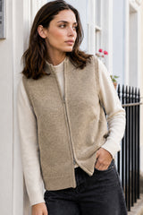 Close-up of a model wearing the Aspiga Merino Wool Zip Through Gilet in Taupe over a cream jumper, leaning against a door