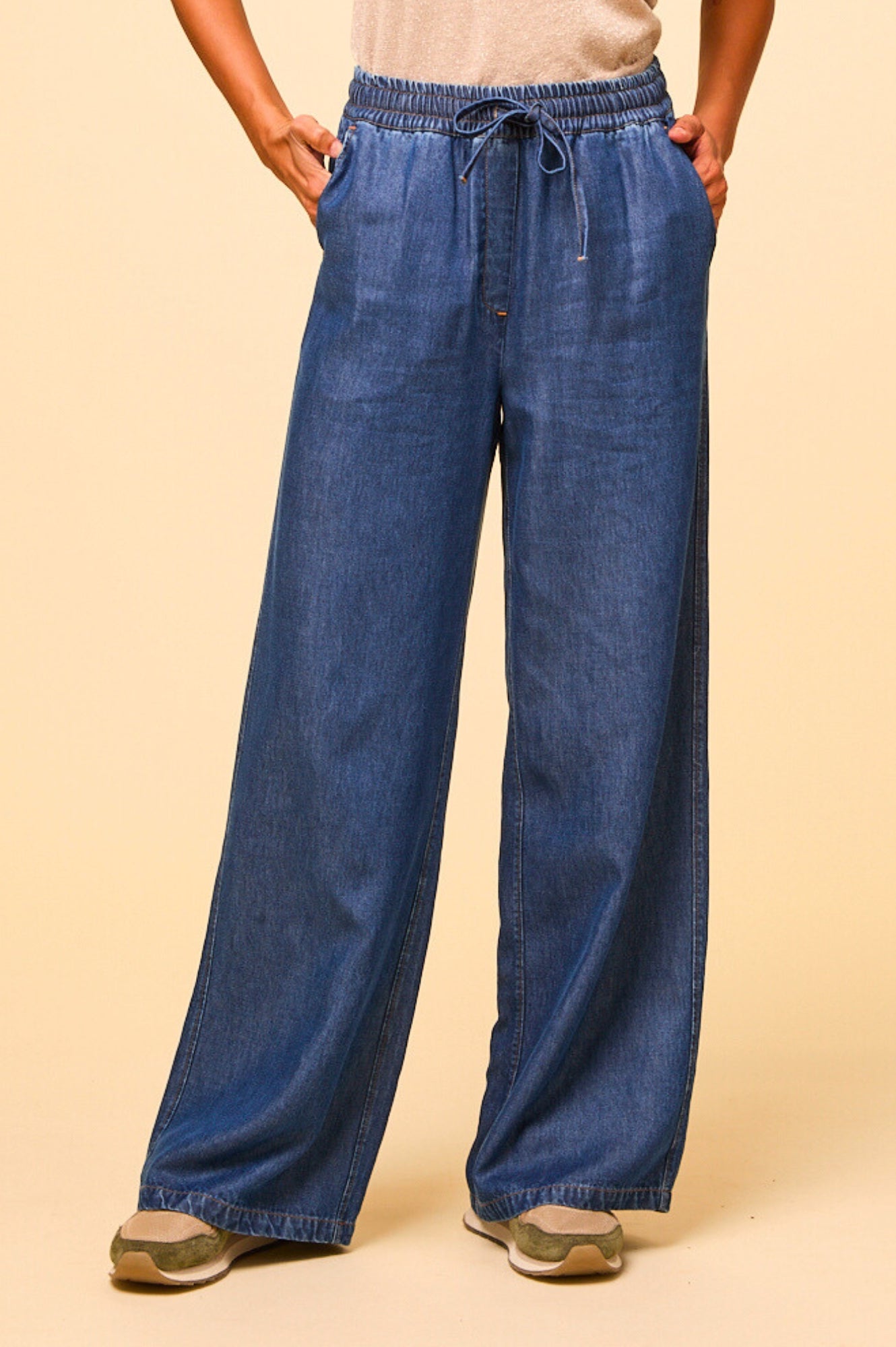 Woman wearing wide-leg denim trousers with drawstring waist and pockets, paired with a beige top and trainers.