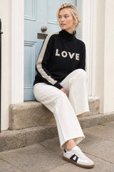 Model wearing the Aspiga Merino Wool Love Polo Neck Jumper in Black/Taupe with a taupe beanie, seated on a wooden bench outdoors with a lighthouse in the background