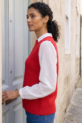 Lifestyle photo of a model wearing the Aspiga Merino Wool Knitted Short Tank in Red, a ribbed knit crew-neck sweater vest layered over a white collared blouse, standing at a doorway