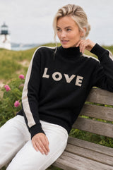Close-up of a smiling model wearing the Aspiga Merino Wool Love Polo Neck Jumper in Black/Taupe with a taupe beanie, seated outdoors, hand near neck