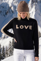 Model wearing the Aspiga Merino Wool Love Polo Neck Jumper in Black/Taupe with a taupe beanie and white trousers, smiling, snowy mountains in the background